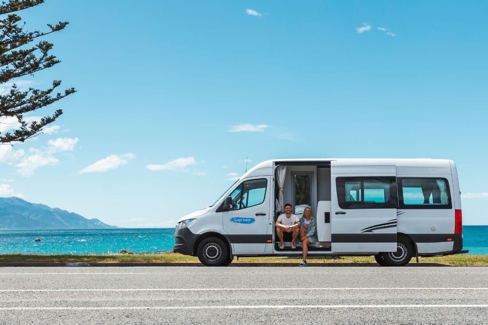 Hvid autocamper parkeret ved kystvej med to personer i døren, hav og bjerge i baggrunden