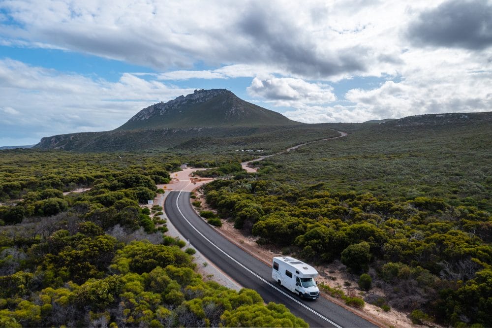 Hvid autocamper kører på en snoet vej gennem grønt busklandskab med et bjerg under skyet himmel