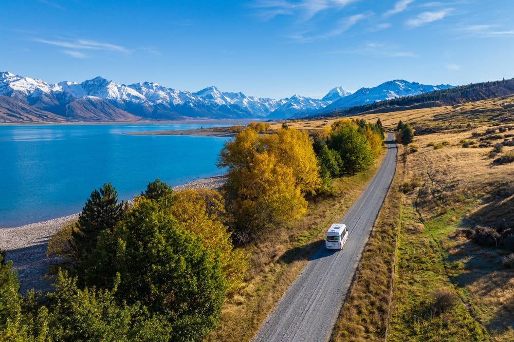 Hvid autocamper kører på en vej langs en turkis sø med sneklædte bjerge i baggrunden