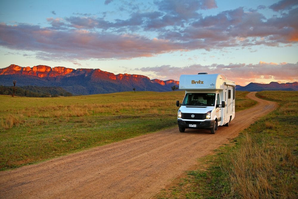 Hvid autocamper på en grusvej gennem åbent græsland med bjerge i solnedgang