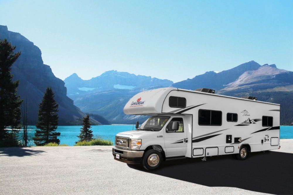 Hvid autocamper parkeret ved en turkis sø med bjerge og fyrretræer i baggrunden