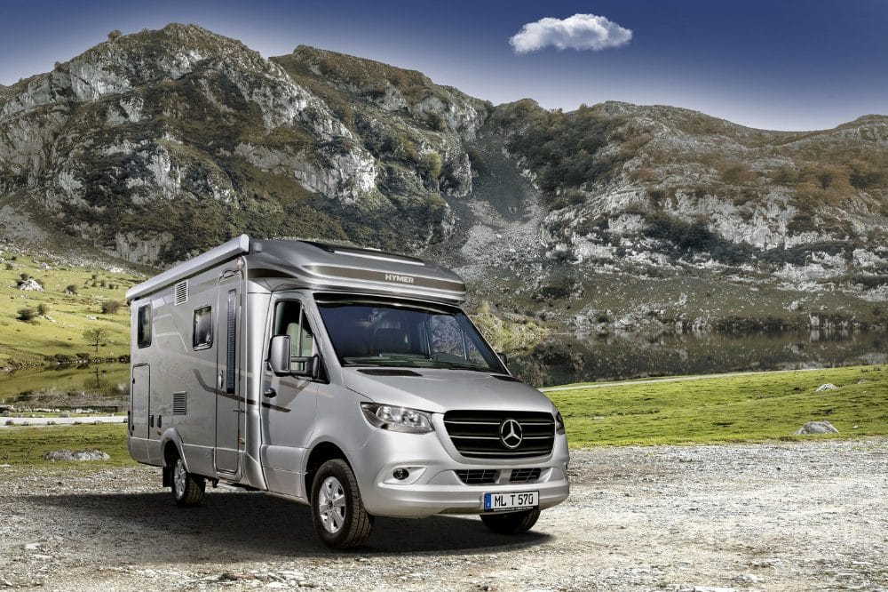 Sølvfarvet autocamper parkeret på grus foran bjerge, grøn eng og en lille sø