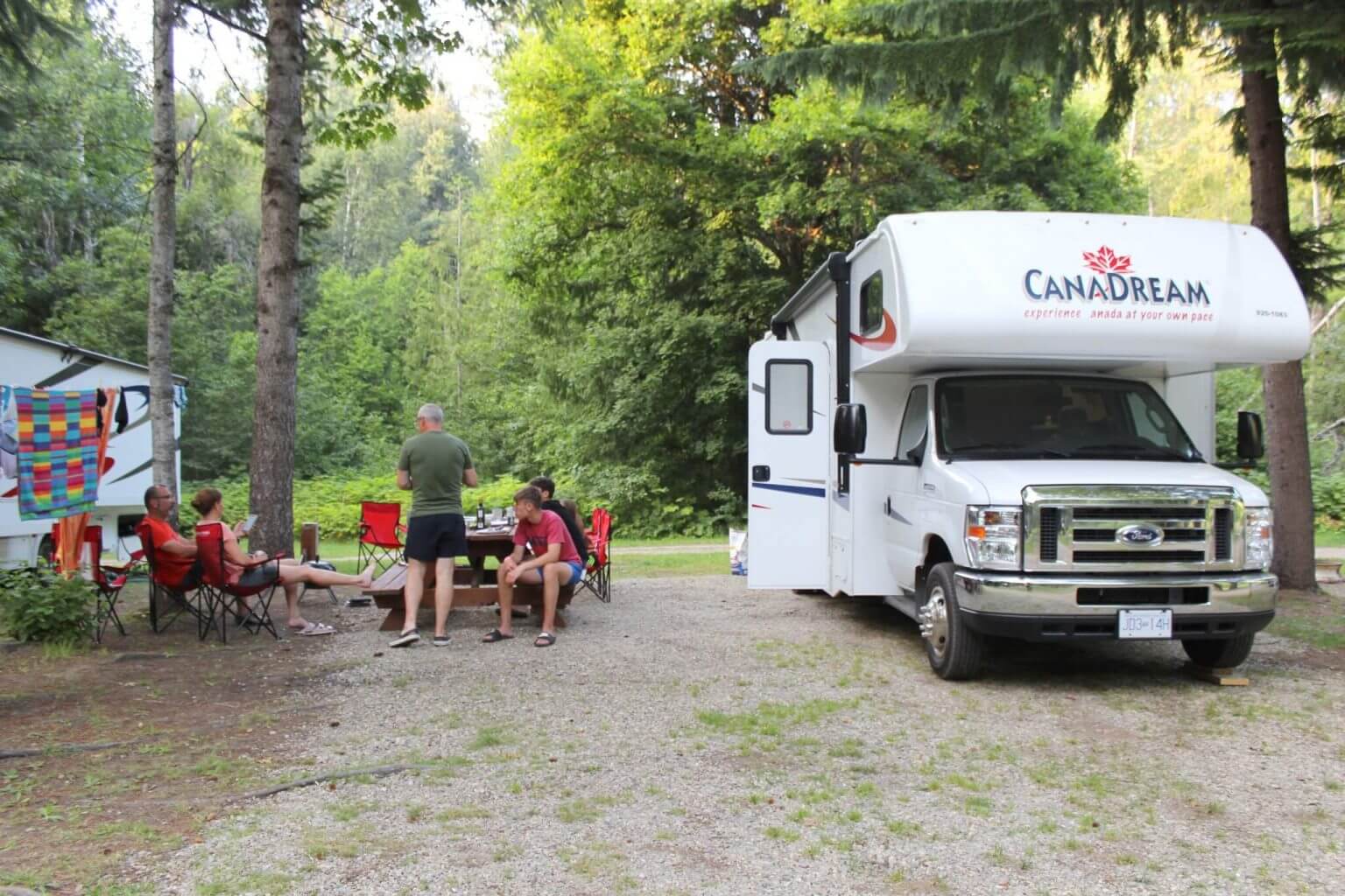 Hvid Canadream autocamper parkeret ved en skovcampingplads med personer omkring et bord